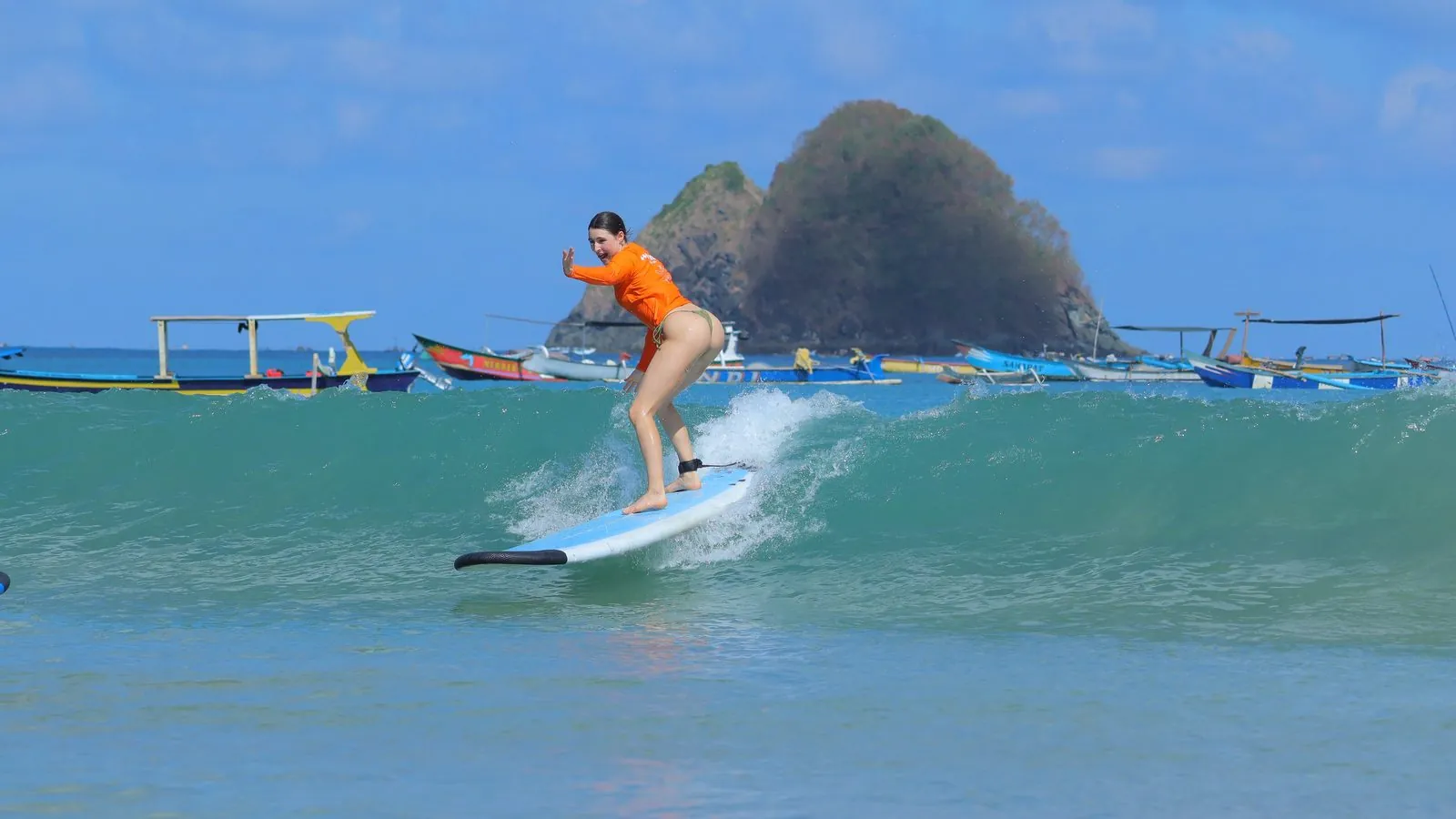 Surfer in an orange Happiness rashguard riding a clean wave past traditional Indonesian boats and a rocky point in Lombok